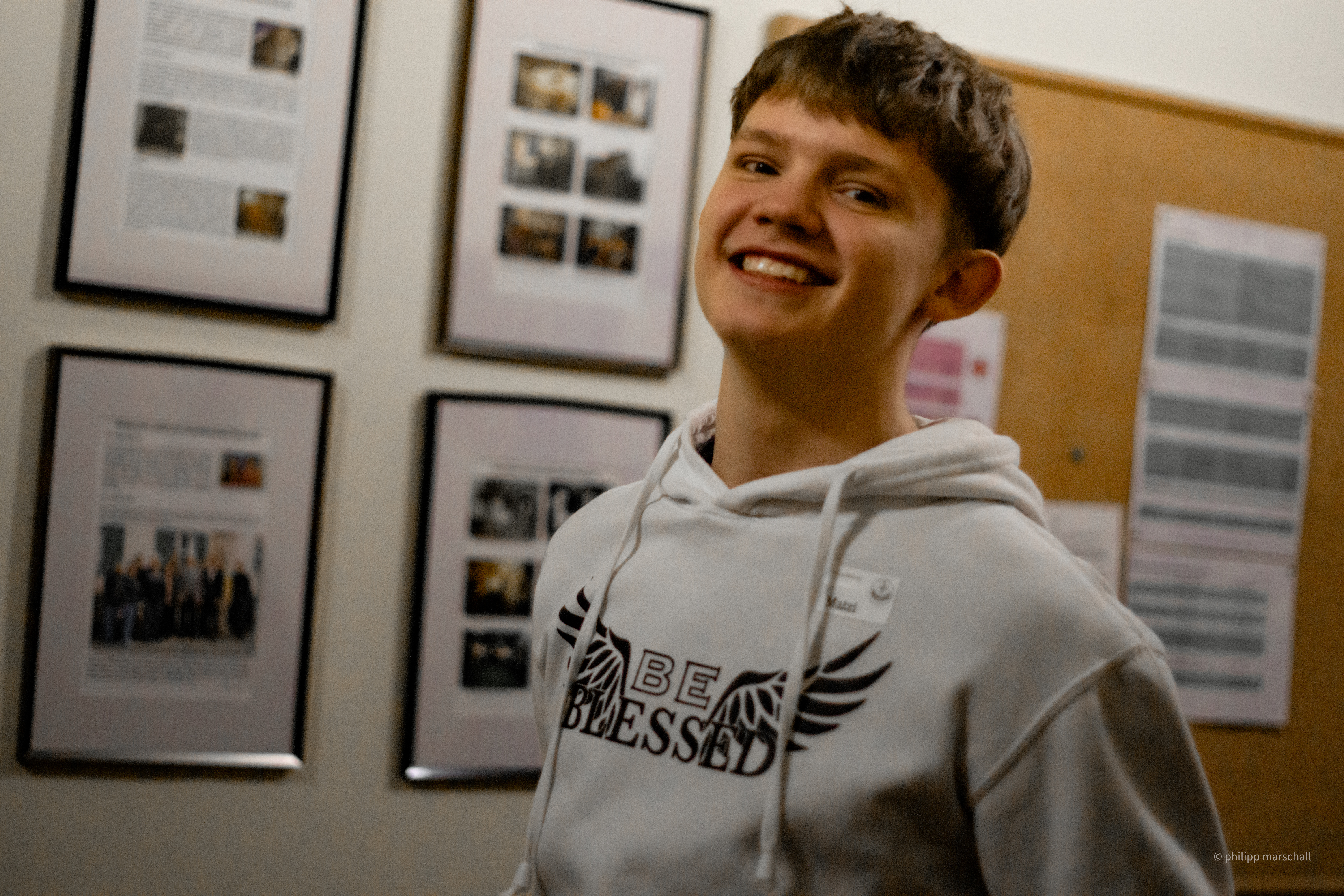Ein lächelnder junger Mann in einem weißen Kapuzenpullover steht vor einer Wand mit gerahmten Fotos und Texten.